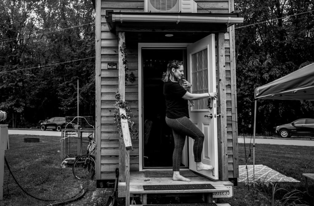Sara steps barefoot out of the door of her tiny house, holding her cat